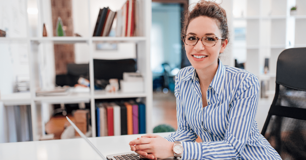 Woman with glasses and striped shirt, hair in a bun, sitting at a desk and looking at the camera